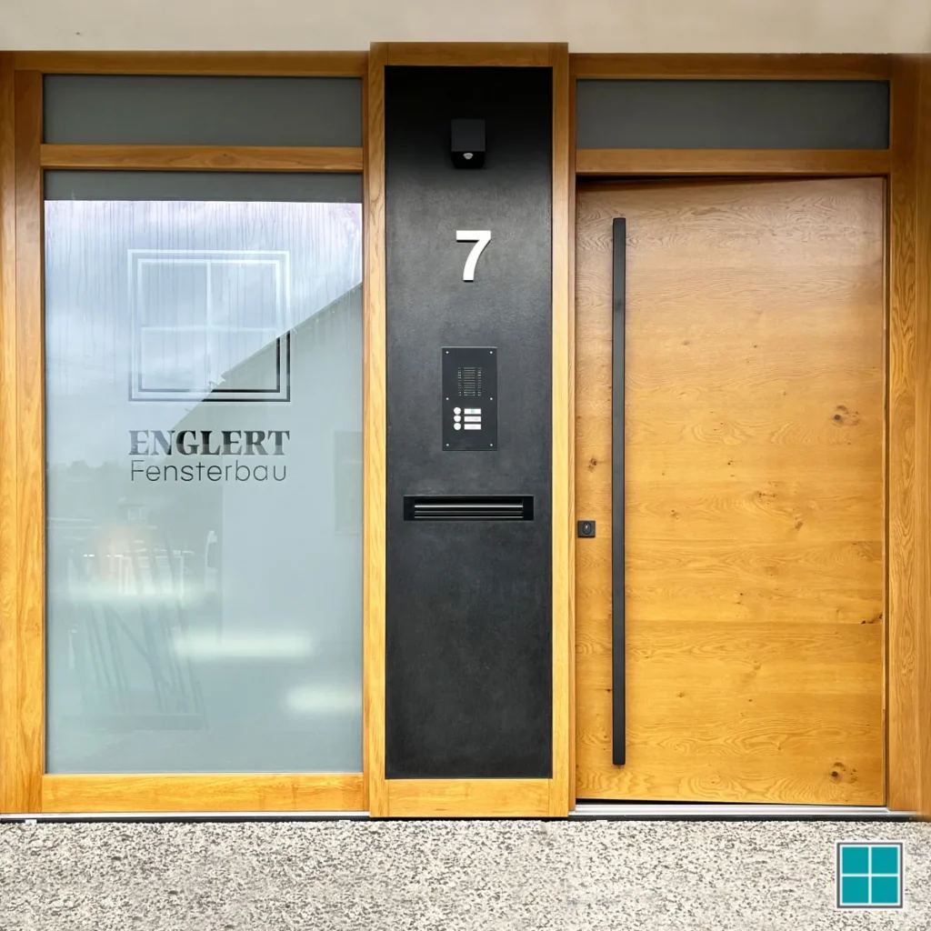 Eingangsbereich von aussen. Man sieht ein bodentiefes Fenster aus Glas und eine hellem Haustüre aus Holz.