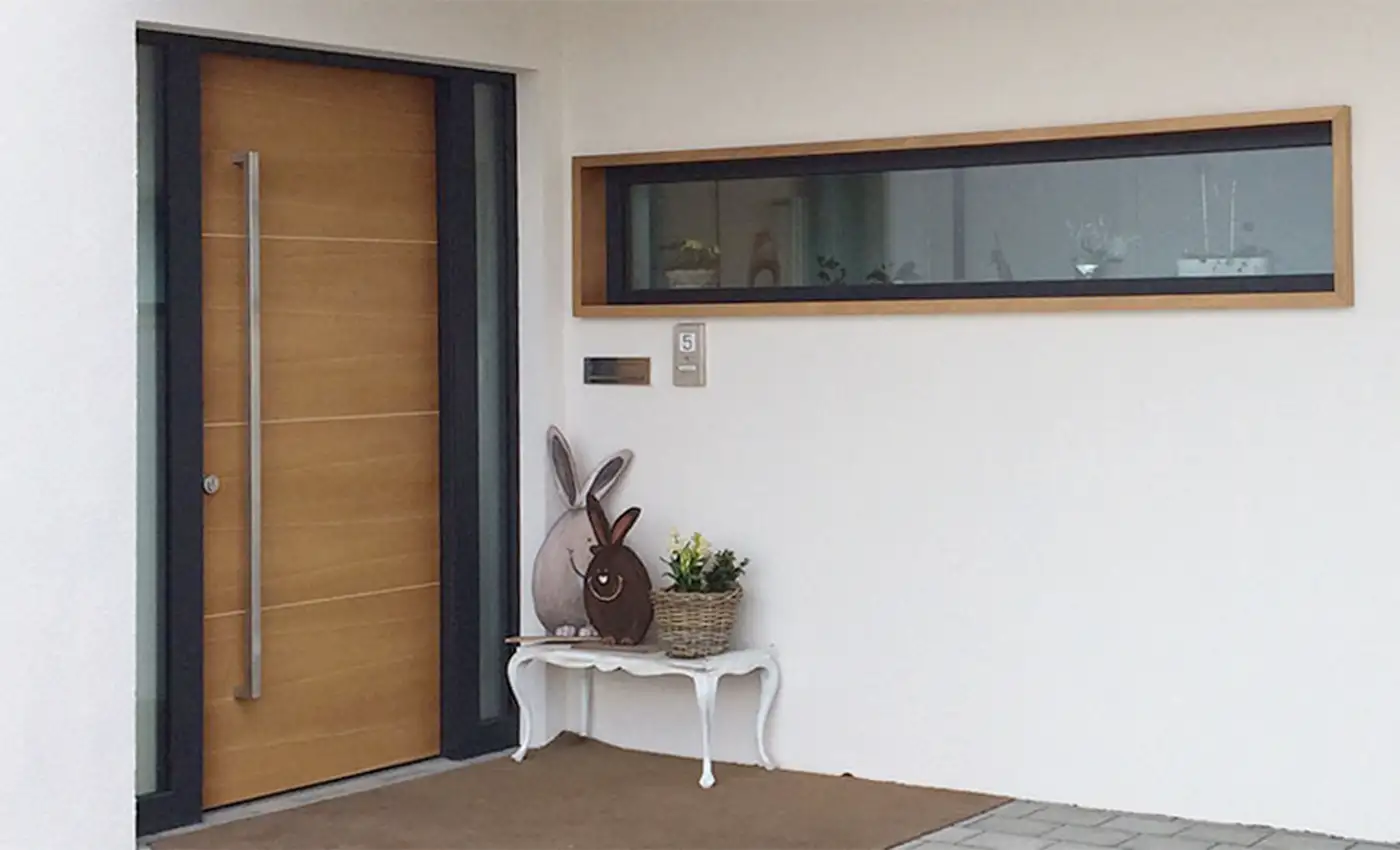 Eingangsbereich eines Hauses mit weißer Fassade, einer holzfarbenen Haustüre und eines Fensters mit Oster-Deko vor der Türe